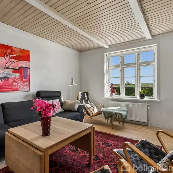 Stue indrettet med sort sofa, træbord med vase med blomster og stol under stort rødt maleri. Vindue med udsigt over landskab. Lyst trægulv og tæppe i røde nuancer.