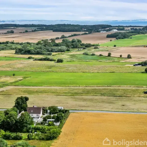 Et hus ligger blandt grønne træer. En traktor arbejder på en mark. Omkring er der åbne landbrugsområder med marker og spredte træer under en blå himmel.