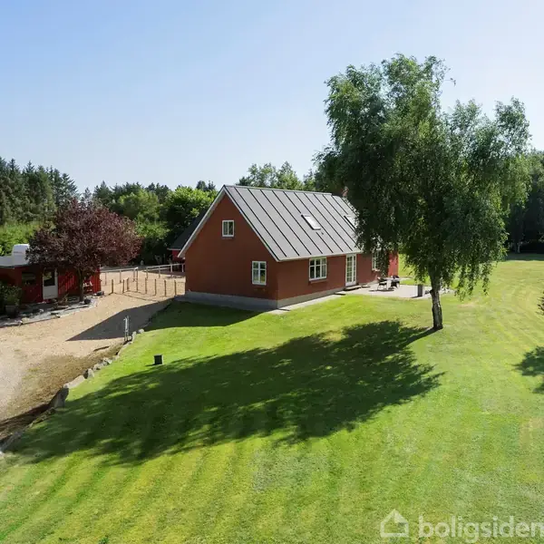 Et rødt hus med sort tag står på en velplejet grøn plæne. Ved siden af findes en grusvej, omgivet af træer og buske under en klar blå himmel.