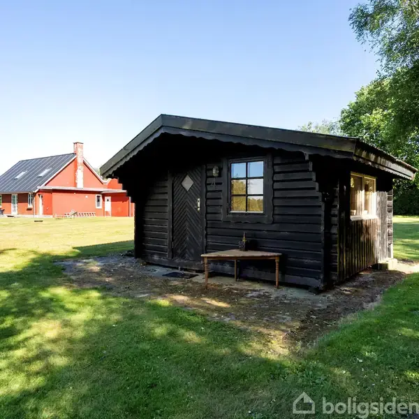 Et sort træskur står på en grøn plæne. I baggrunden ses et rødt hus med solpaneler på taget, omringet af træer og blå himmel.