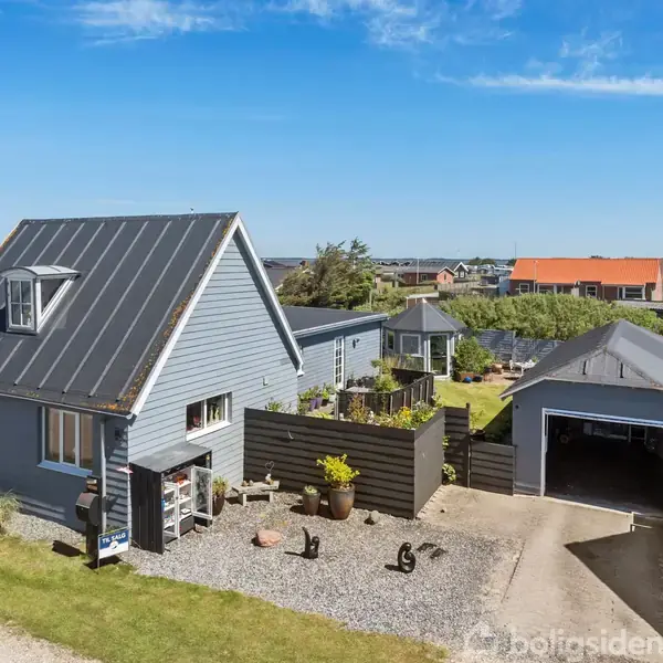 Et gråt hus med skråt tag står ved en grussti. Ved siden af ses en garage. Haven har grønne planter og hegn. Blå himmel og flere huse i baggrunden.