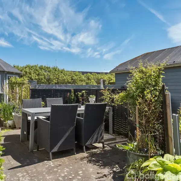 Terrassemøbler står på en solrig terrasse omgivet af planter. En lyseblå bygning til venstre og en større bygning til højre omkranser området. Himmel er klar med få skyer.