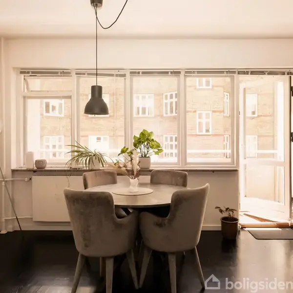 Et rundt bord med grå polstrede stole står i centrum af en lys spisestue, omgivet af store vinduer, en pendellampe og indendørs planter, der giver et moderne, hyggeligt udtryk.