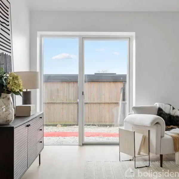 Stue med en lys lænestol ved siden af en gulv-til-loft-terrassedør, der fører ud til en have. En kommode med vase og lampe står ved siden af.