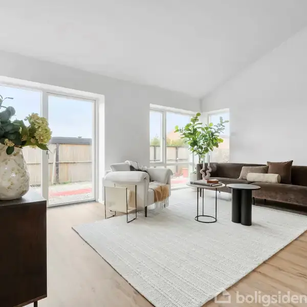 Stue med moderne indretning; en brun sofa står på et hvidt tæppe ved siden af et lille sofabord, omgivet af store vinduer, der giver naturligt lys.