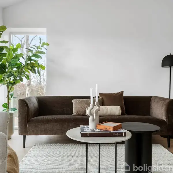 En mørkebrun sofa står i stuen ved siden af et stort vindue. En stueplante og en sort gulvlampe er i nærheden. Foran står to runde borde med bøger og lys.