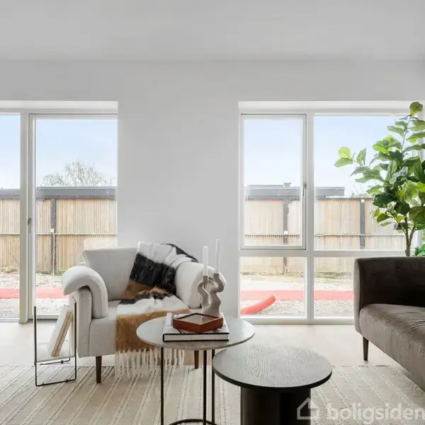 Lyst stue med to glasdøre, stilfuldt møbleret med en hvid stol og brun sofa. Grøn plante i hjørnet, små borde foran, udsigt til terrasse gennem vinduerne.