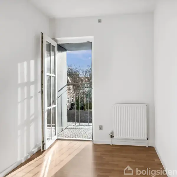 Et lyst, tomt rum med trægulv og en åben dør fører ud til en lille balkon. Radiator står ved væggen under et vindue.