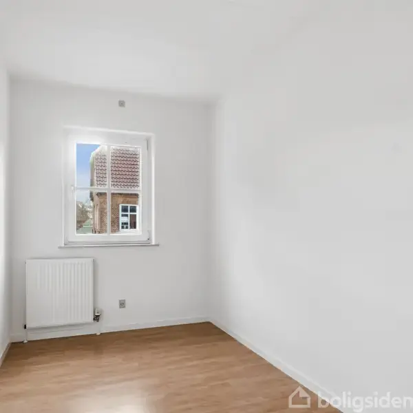 Et tomt værelse har en hvidmalet væg og trægulv. Et vindue lader dagslys strømme ind og viser et hustag udenfor. En radiator er placeret under vinduet.
