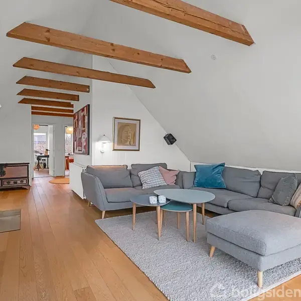 Et gråt hjørnesofa-sæt står på et tæppe, ved siden af et rundt sofabord og en standerlampe. I baggrunden ses en brændeovn ved en væg med synlige bjælker.