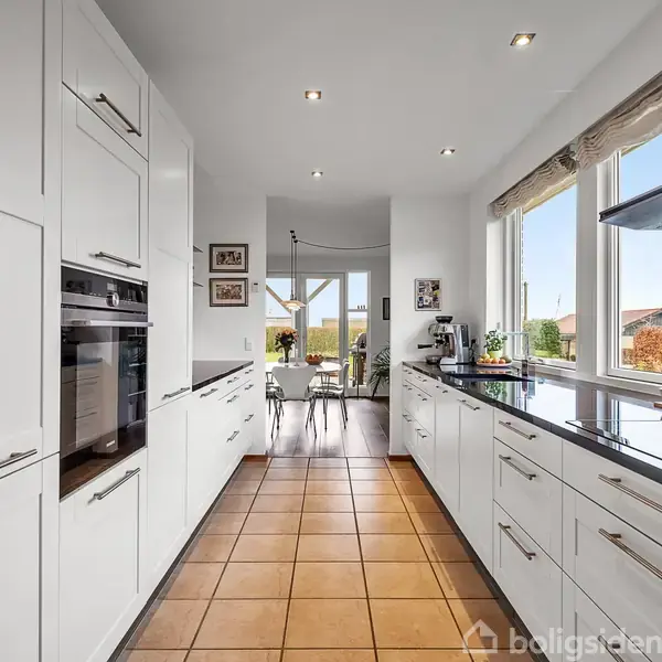 Hvidt køkken med sorte bordplader har indbyggede apparater og store vinduer, der giver udsigt til en solrig terrasse og have. Flisegulv fører til spiseområde med glasdøre.