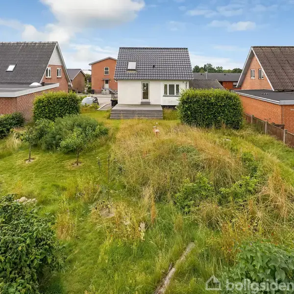 Et hvidt hus med grått tag står i en have med højt græs, omgivet af nabohuse med røde mursten. En træterrasse ses foran huset.