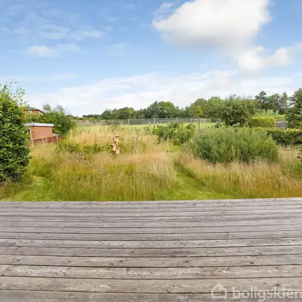 En træterrasse strækker sig ud mod en have med højt græs og buske. I baggrunden kan ses et åbent grønt område med træer og blå himmel med skyer.