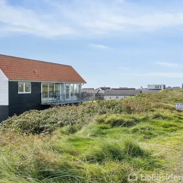Et moderne hus med rødt tag og glasværn står roligt. Det ligger højt i et kuperet, grønt landskab med en sti i nærheden. Et skilt i græsset ses tydeligt.