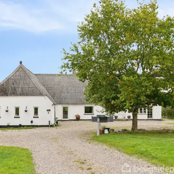 En hvid landbygning med skråt tag ligger ved en grusvej, omgivet af en grøn græsplæne og et stort træ. Marker kan ses i baggrunden under en blå himmel.