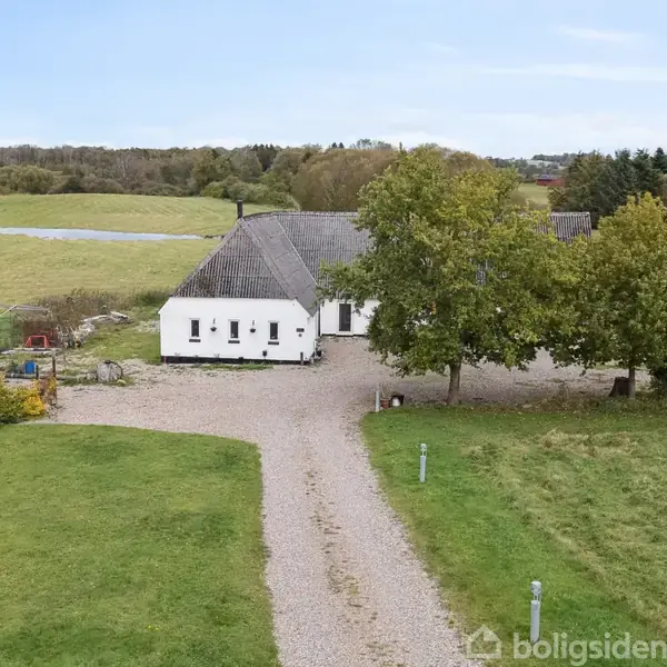 En hvid gårdbygning står stille omgivet af grønne marker og træer. En grusvej fører op til indgangen, mens en lille hytte kan ses til venstre i billedet.
