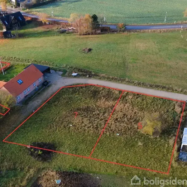 En åben grund omkranset af røde linjer viser en byggegrund med to huse og en grusvej i et landligt område, omgivet af marker og spredt bebyggelse.