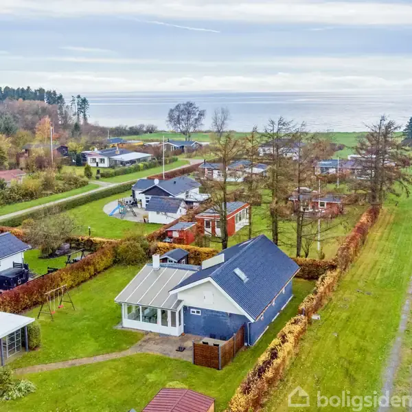 Huse omgivet af grønne haver ved en grusvej tæt på en kystlinje. Træer og buske adskiller ejendommene, mens havet kan ses i baggrunden under en overskyet himmel.