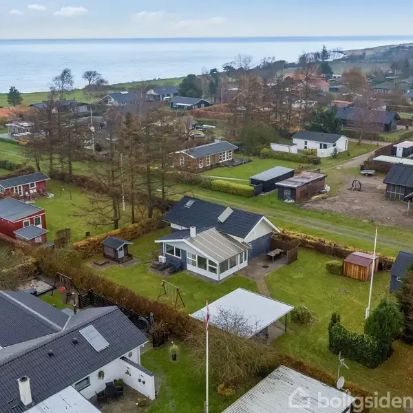 Huse set oppefra står stille blandt grønne haver nær kysten. Området omfatter forskellige ejendomme med græsplæner og træer. Havet ses i baggrunden under en let overskyet himmel.