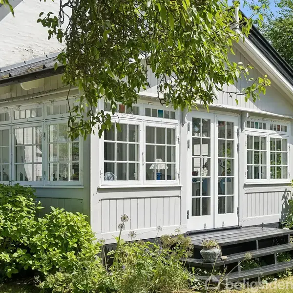 Et lille, hvidt hus med store vinduer og en træterrasse foran. Omgivet af grønne træer og buske i en frodig have.