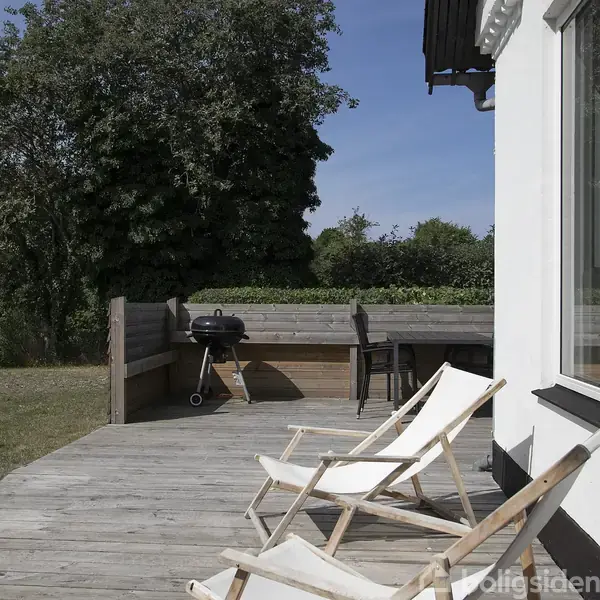 To liggestole står på en træterrasse foran et hus. En grill og et bord med stole er i baggrunden, omgivet af grønne træer og en græsplæne.