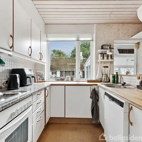 Køkken med hvide skabe og træbordplade. På bordet står kaffemaskine og elkedel. Et vindue over vasken giver udsigt til en have. Flere køkkenredskaber ses i baggrunden.