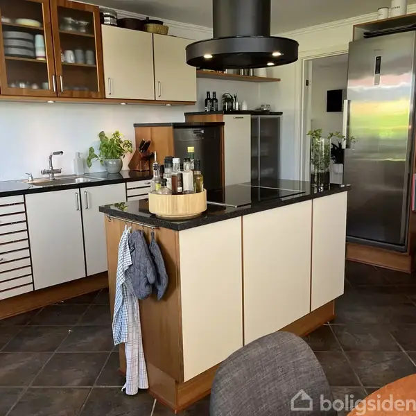 Køkken med hvidt bord og køkkenø, hvor der står flasker og krydderier. Omkring er der skabe, køleskab og hylder med bøger og planter i et moderne rum.