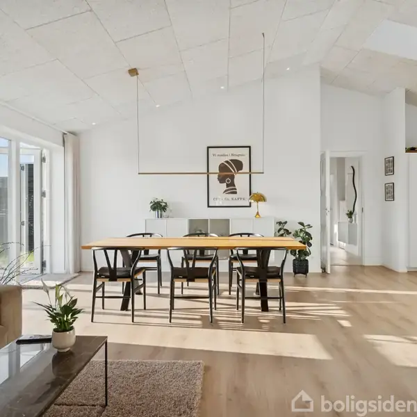 Spisebord med seks stole står i et lyst rum med hvide vægge og trælaminatgulv. Store vinduer slipper sollys ind, mens planter og vægkunst skaber en hyggelig atmosfære.