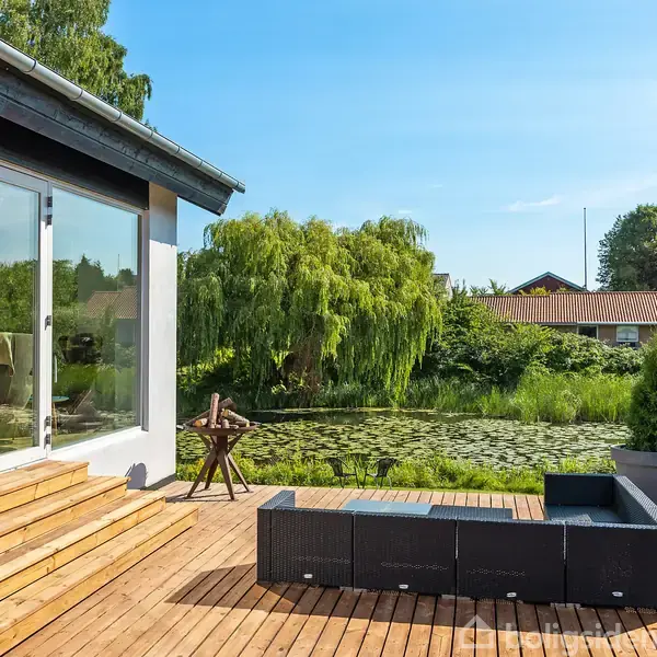 En moderne terrasse med sorte havemøbler står foran en bygning med glasdøre. Terrassen har udsigt over en dam omgivet af grønne træer og buske under en klar blå himmel.