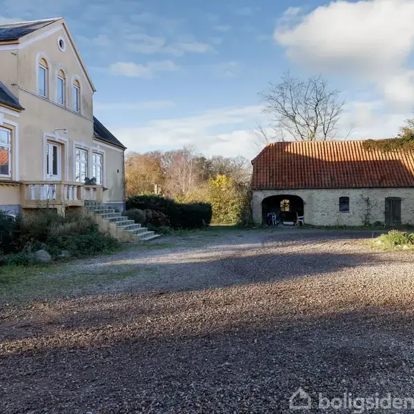 En lys gul bygning med vinduer står til venstre, omgivet af en gårdsplads med grus og en garage eller staldbygning til højre. Baggrunden har træer og en blå himmel.