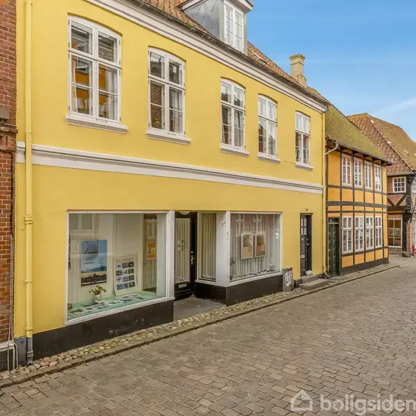 En gul, to-etagers bygning med store vinduer står stille langs en brostensbelagt gade i en række af historiske huse, der strækker sig mod horisonten.