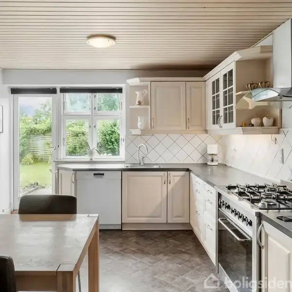 Et køkken med hvidt skab og grå bordplade, stor gaskomfur og emhætte. Et spisebord med skål står i midten. Et vindue viser en grøn have udenfor.