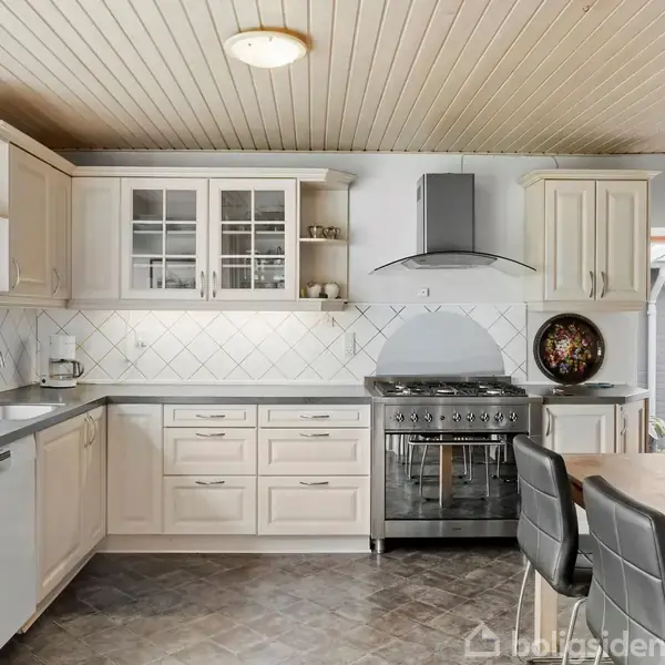 Et moderne køkken har lyse skabe, en indbygget ovn med emhætte, og et spisebord med stole. Væggene er flisebelagte, og rummet har et lyst trægulvloft.