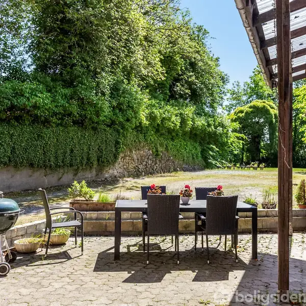 Grill og havemøbler står stille på en flisebelagt terrasse ved siden af et rødt murstenshus. Terrassen er omkranset af grønne buske og store træer.