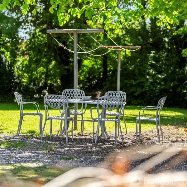 Et metalbord med fire stole står på en grusplads i en have, omgivet af frodig, grøn vegetation og træer, i sollys.