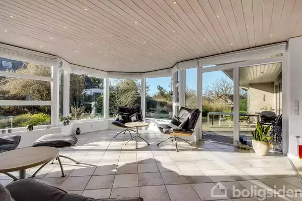 En stue med store panoramavinduer, sorte lænestole og sofabord. Rummet er lyst og åbner ud til en terrasse, omkranset af en frodig have.