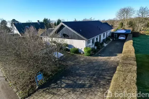 En hvid villa med sort tag står langs en flisebelagt indkørsel. En bil er parket under en carport. Omgivet af buske og træer i et beboelsesområde.