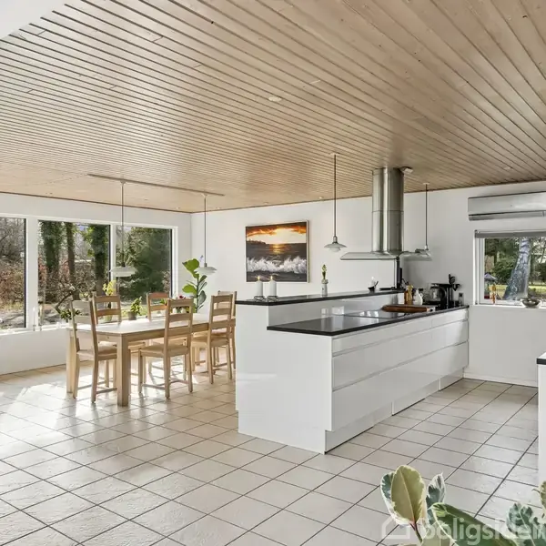 Et lyst køkken med en stor ø med sort bordplade. Langbord med stole ved vinduer. Træloft og flisegulv. Maleri på væggen og udsigt til have.
