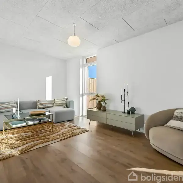 En udendørs terrasse med flettemøbler står ved siden af en moderne murstensbygning. I baggrunden ses en carport, en træbeklædt skur og flere campingvogne på en byggeplads.