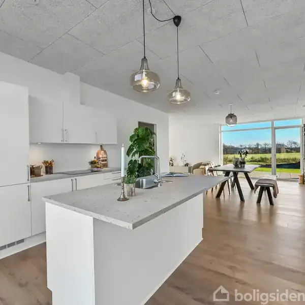 Et lyst køkken med hvidt interiør har en central ø med planter og lamper over. I baggrunden er der et spiseområde med udsigt til en grøn have gennem store vinduer.