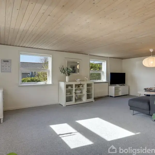 Stue med gråt hjørnesofa, lavt tv-bord og hvid skænk med dekorationer. To vinduer giver naturligt lys. Loft er i lyst træ, og en stor lampe hænger over sofaen.