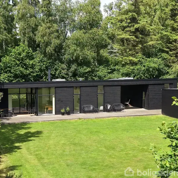 Sort, moderne enetageshus står på en grøn græsplæne, omgivet af høje, frodige træer. En terrasse med havemøbler foran husets store glaspartier.