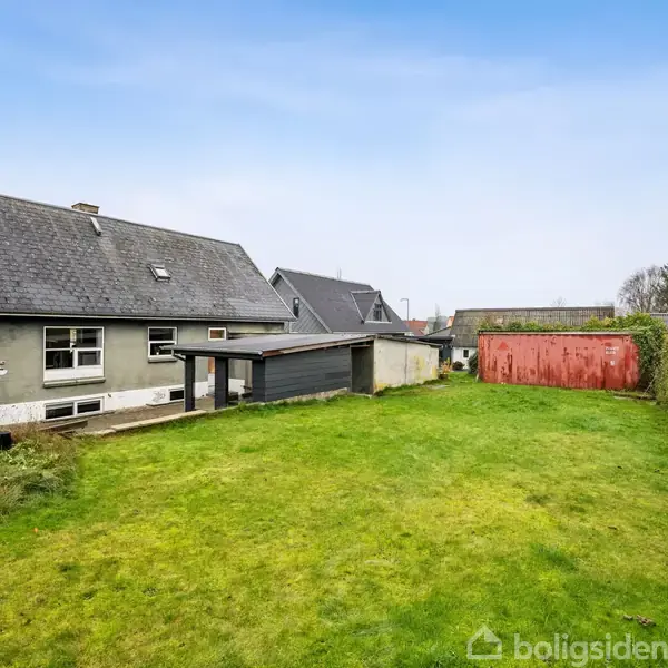 Hus med gråt tag og vinduer står stille ved siden af garage i en græsplænehave, omgivet af hække og et rødt skur.