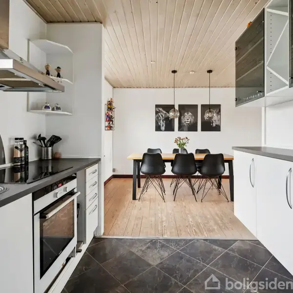 Køkken i hvidt design har ovn og emhætte. En åbenhylde over køkkenbordet. I baggrunden ses en spiseplads med et træbord og sorte stole, lys træloft og vægkunst.