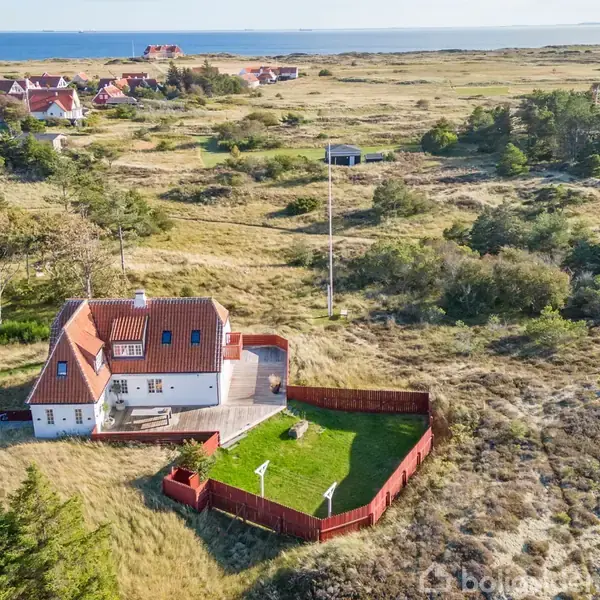 Hus med rødt tag og hvid facade omgivet af en grøn have med rødmalet hegn. Beliggende i et åbent, grønt landskab nær en kystby og havet i baggrunden.