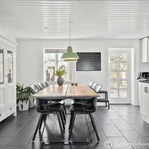 Et spisebord i træ er omkranset af sorte stole i en lys spisestue. En grøn pendellampe hænger over bordet. Vinduerne giver udsigt til træer udenfor.