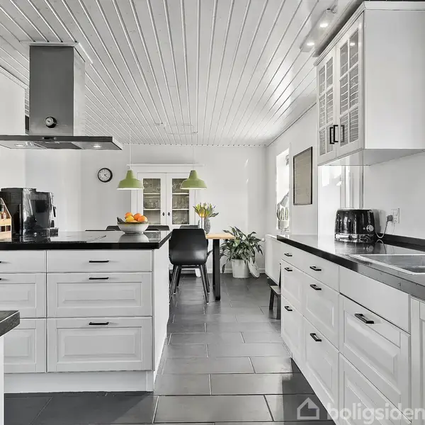 Et moderne køkken med hvide skabe, sort bordplade og stålvask; på køkkenøen står en brødrister og frugtskål. I baggrunden ses et spisebord med grønne lamper.