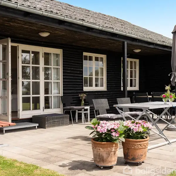 Et sort træhus med overdækket terrasse. Terrassebord og stole står på fliser, omgivet af blomsterkrukker. Grøn have og blå himmel ses i baggrunden.