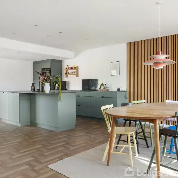 Et rummeligt køkken har en grøn køkkenø og sideborde. Rundt spisebord med farverige stole ses foran en væg med lodrette træpaneler. Et lyst, minimalistisk design dominerer rummet.