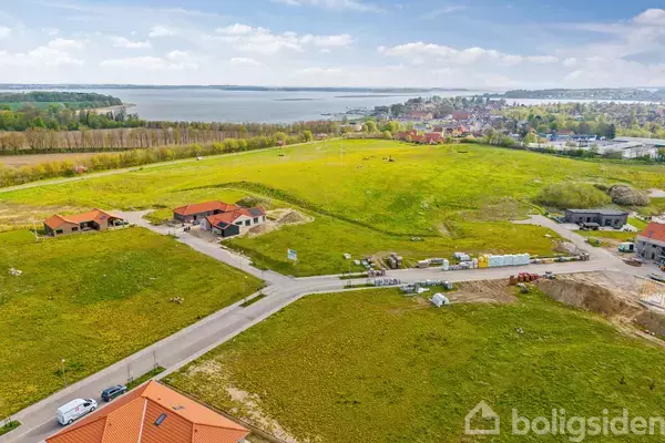 Et åbent grønt landskab med vej, hvor der bygges flere huse med røde tage. I baggrunden ses en kystlinje og en by med træer og røde tage.